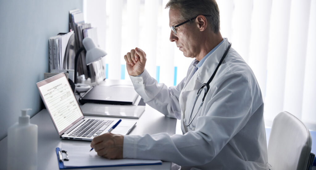 Doctor mayor serio usando una computadora portátil en la consulta médica.