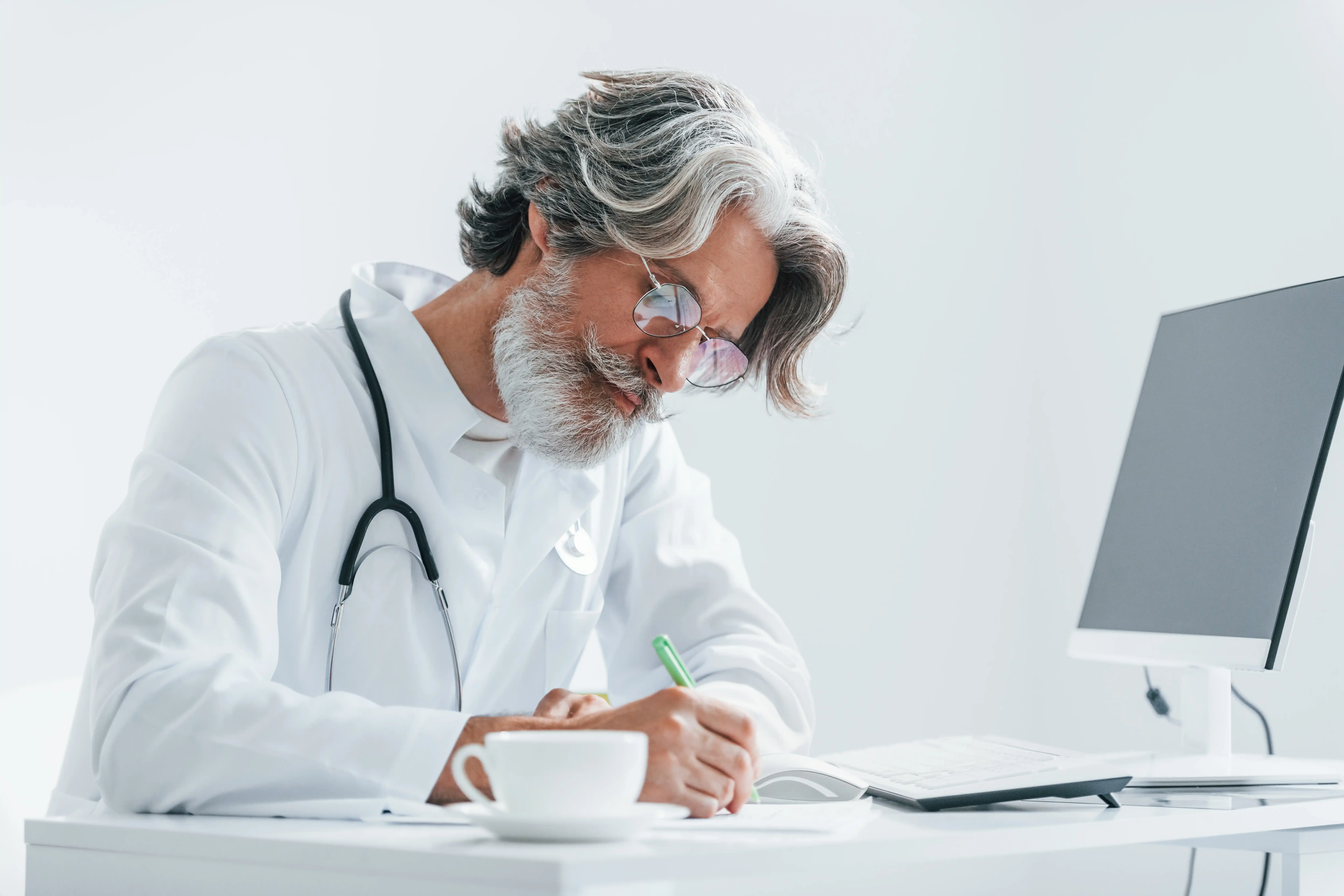 Escribiendo en la mesa del ordenador. Médico mayor con cabello y barba canosos, en bata blanca, dentro de la clínica.